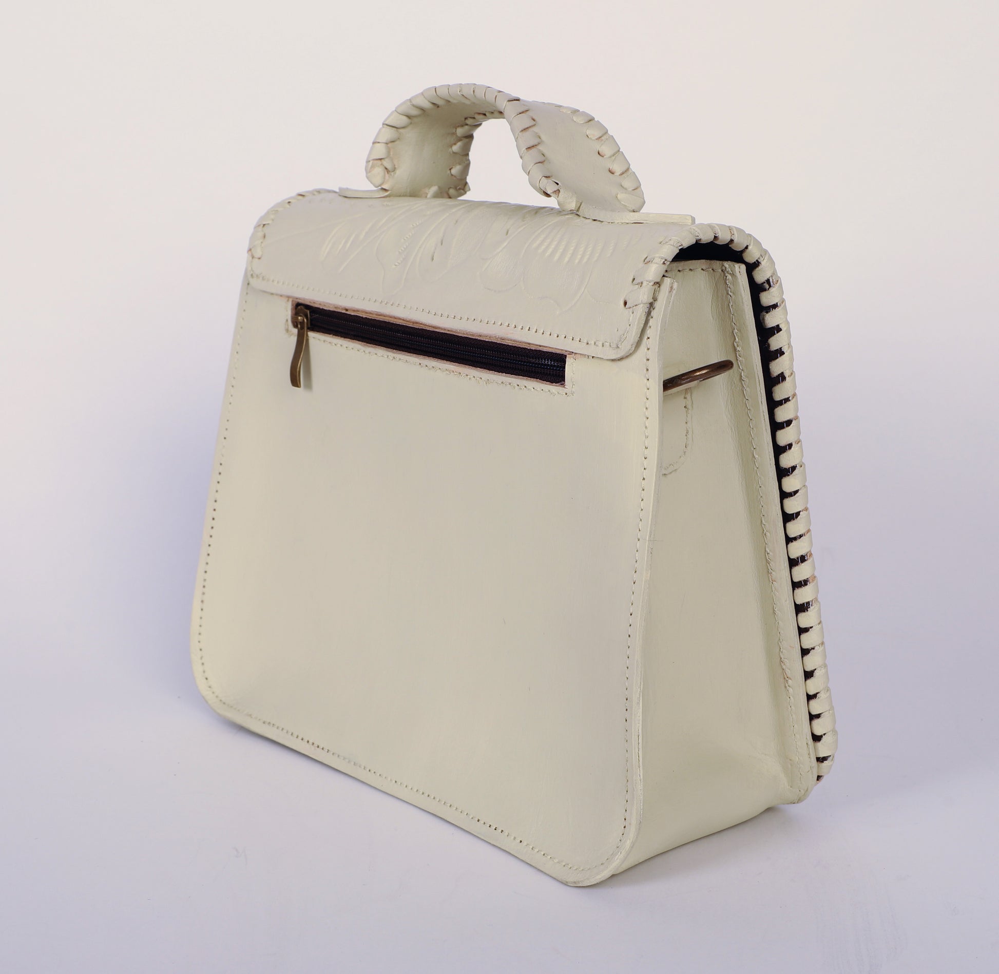 Medium-sized authentic leather handbag with a traditional mexican etching design on the upper flap. Braided handle on top. Fully lined with suede.  Back side view showing zipper and hook to hang crossbody strap. In Color white.